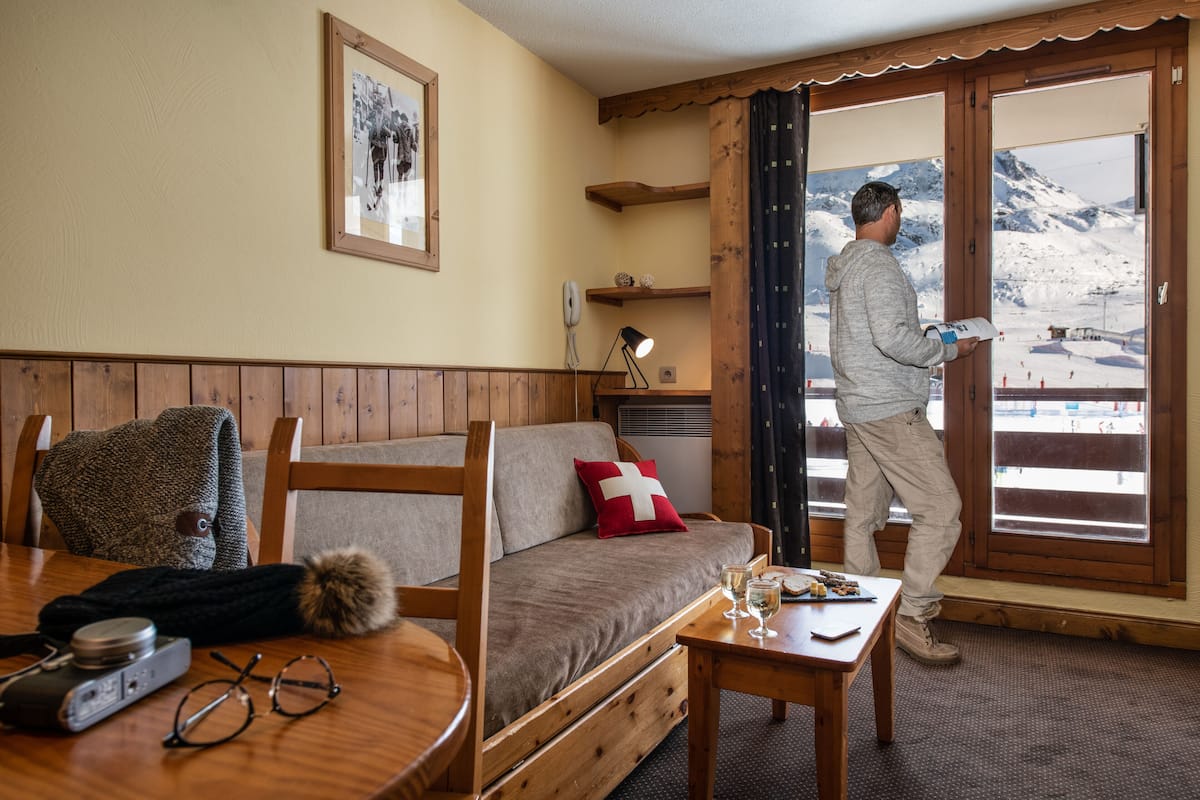 Una habitación acogedora con un sofá de madera, una mesa con una cámara y vasos, y una ventana con vistas a una montaña nevada.