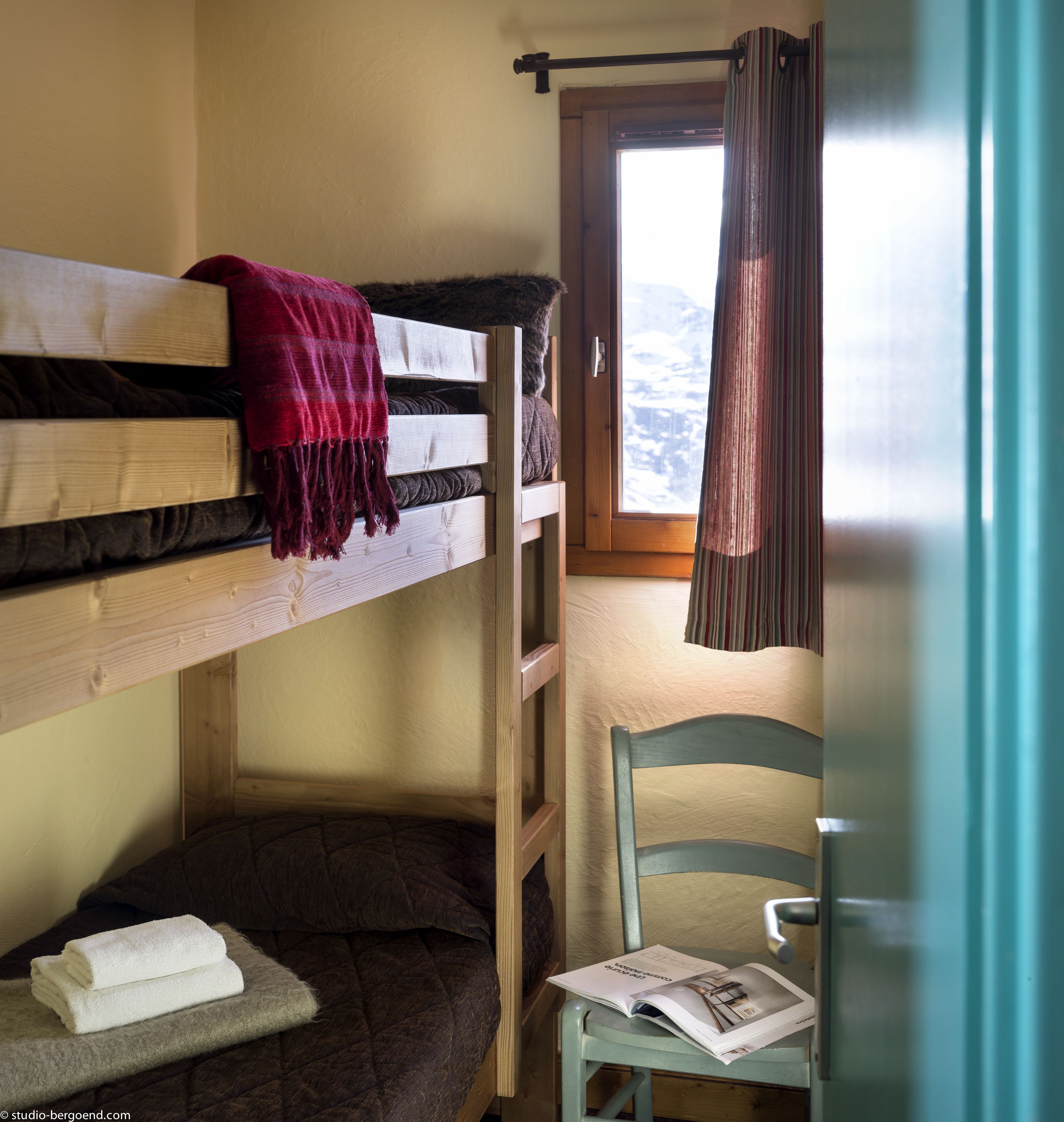 A bunk bed with a red blanket, a chair with a magazine, and a window with curtains.