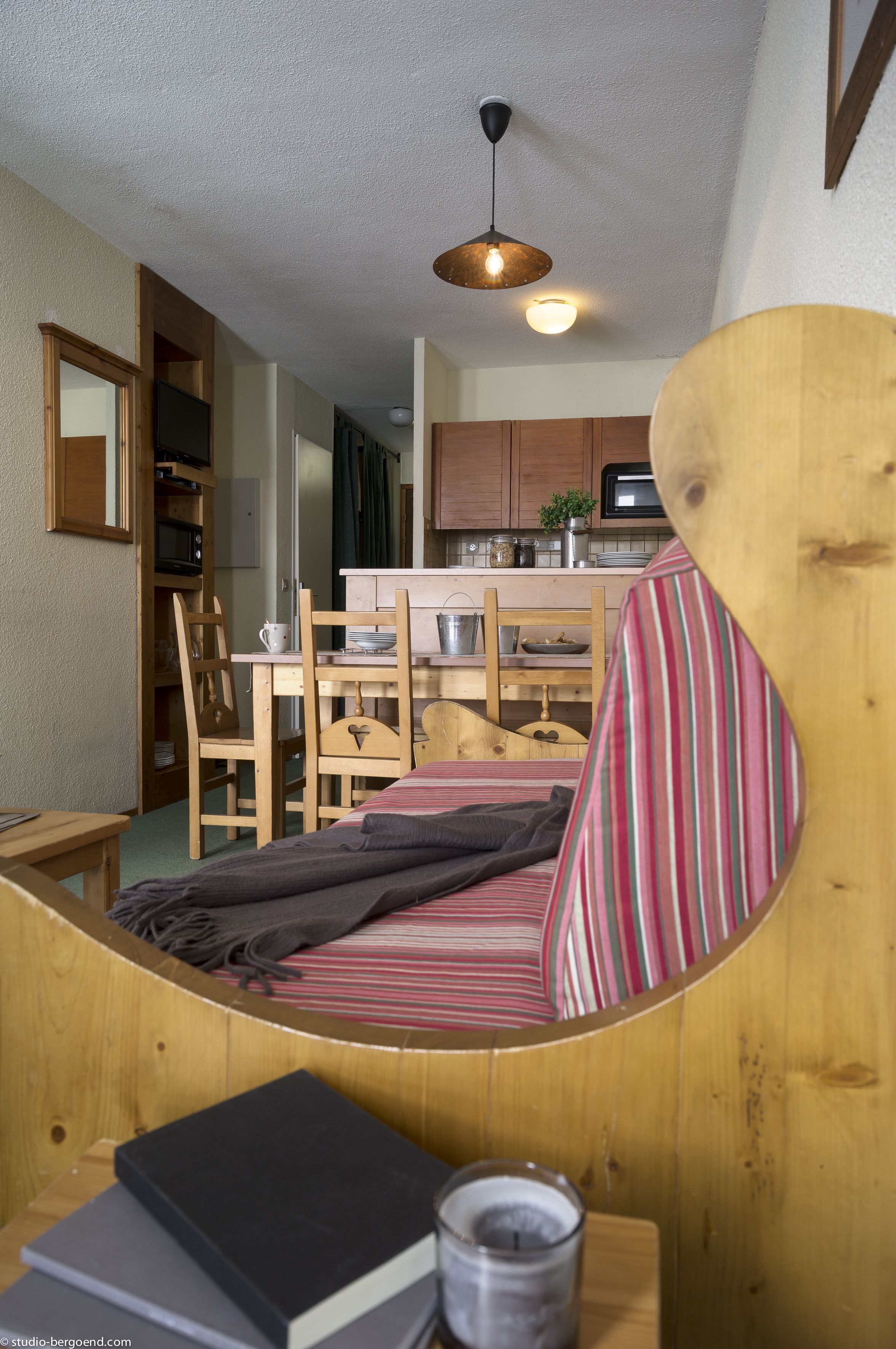 Une pièce chaleureuse avec un lit, une table en bois sur laquelle sont posés des livres, une lampe, et une vue sur la cuisine et la salle à manger.