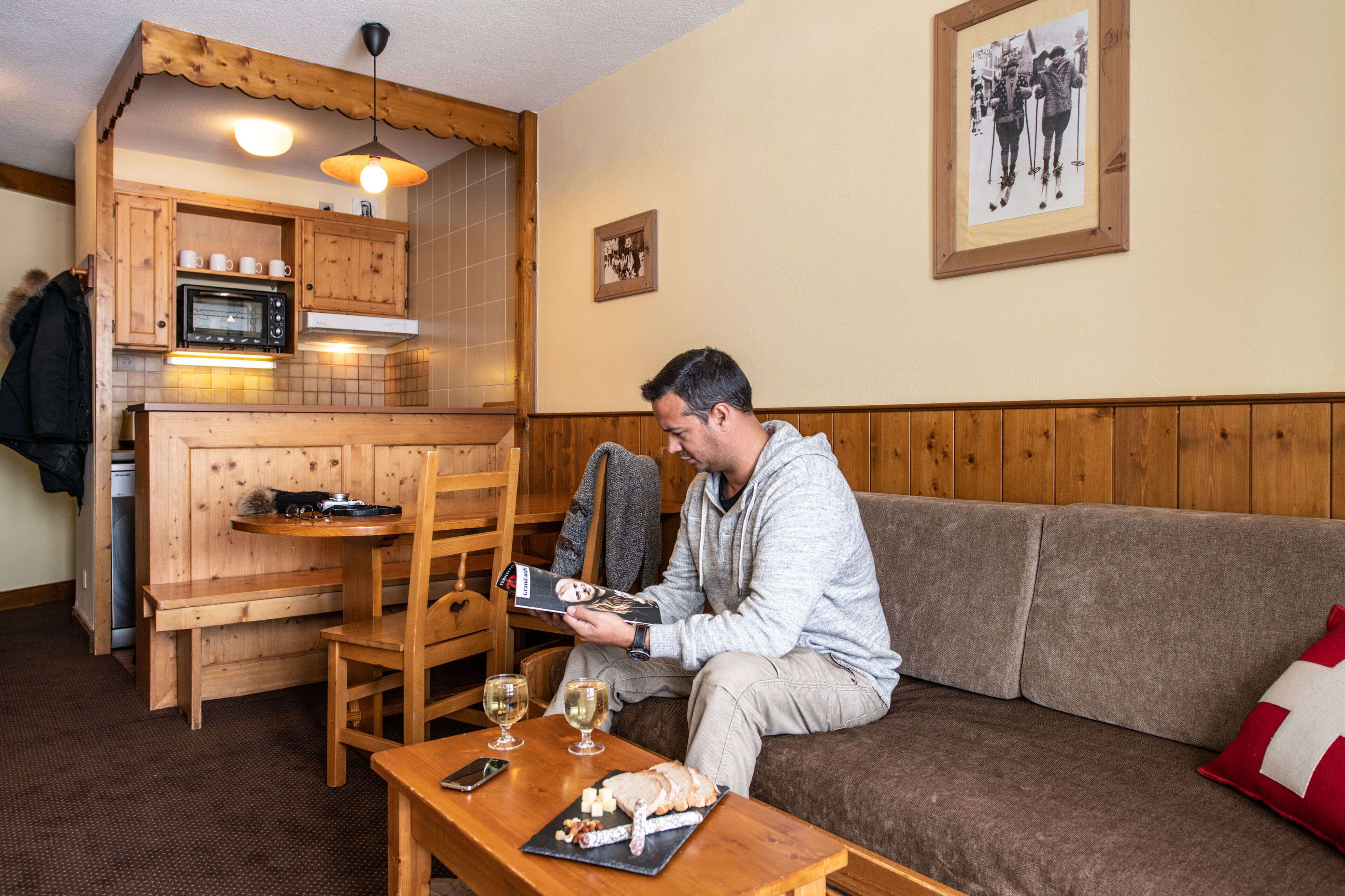A cozy living area with a wooden sofa, a small wooden table with a plate of food and glasses, and a microwave in the background.