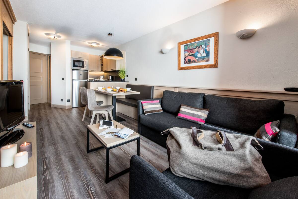 Un salon moderne avec un canapé, une table basse et un téléviseur. On aperçoit à l’arrière-plan un coin cuisine équipé d’un four à micro-ondes et d’une table à manger.