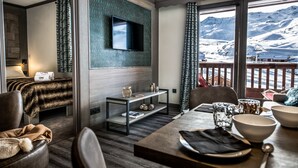 A modern hotel room with a wooden floor, a flat-screen TV mounted on the wall, a dining table with bowls and a spoon, and a view of snow-covered mountains through large windows. - Residence Village Montana (Les Belleville)