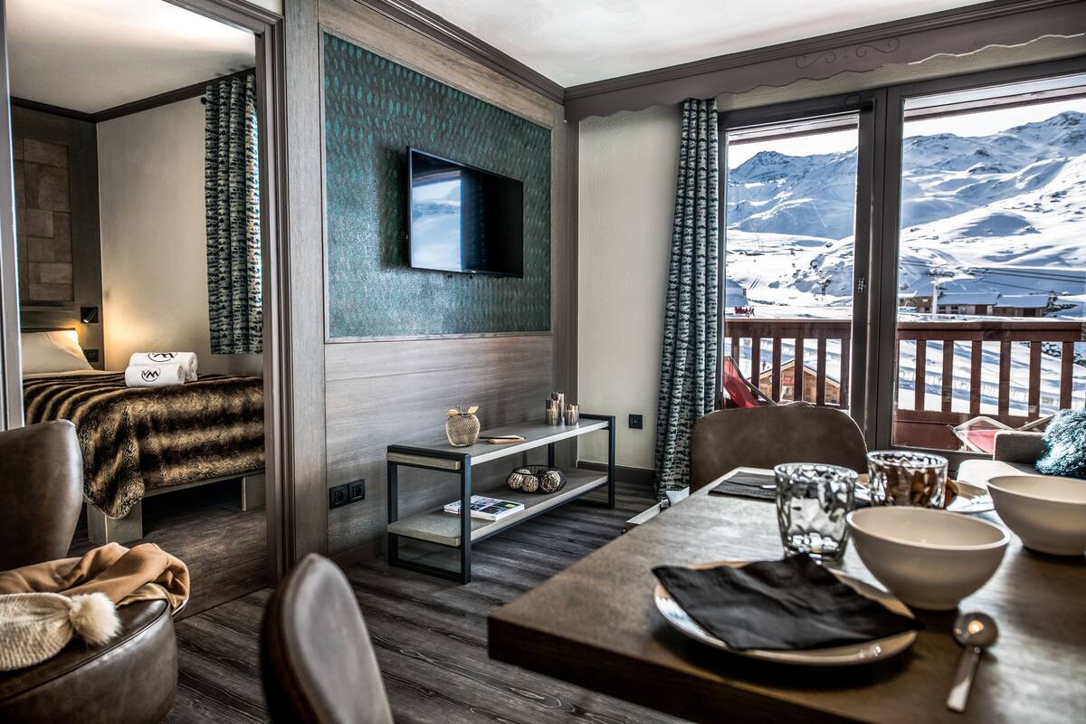 Une chambre d’hôtel moderne avec un plancher en bois, un téléviseur à écran plat fixé au mur, une table à manger avec des bols et une cuillère, et une vue sur des montagnes enneigées par de grandes fenêtres.