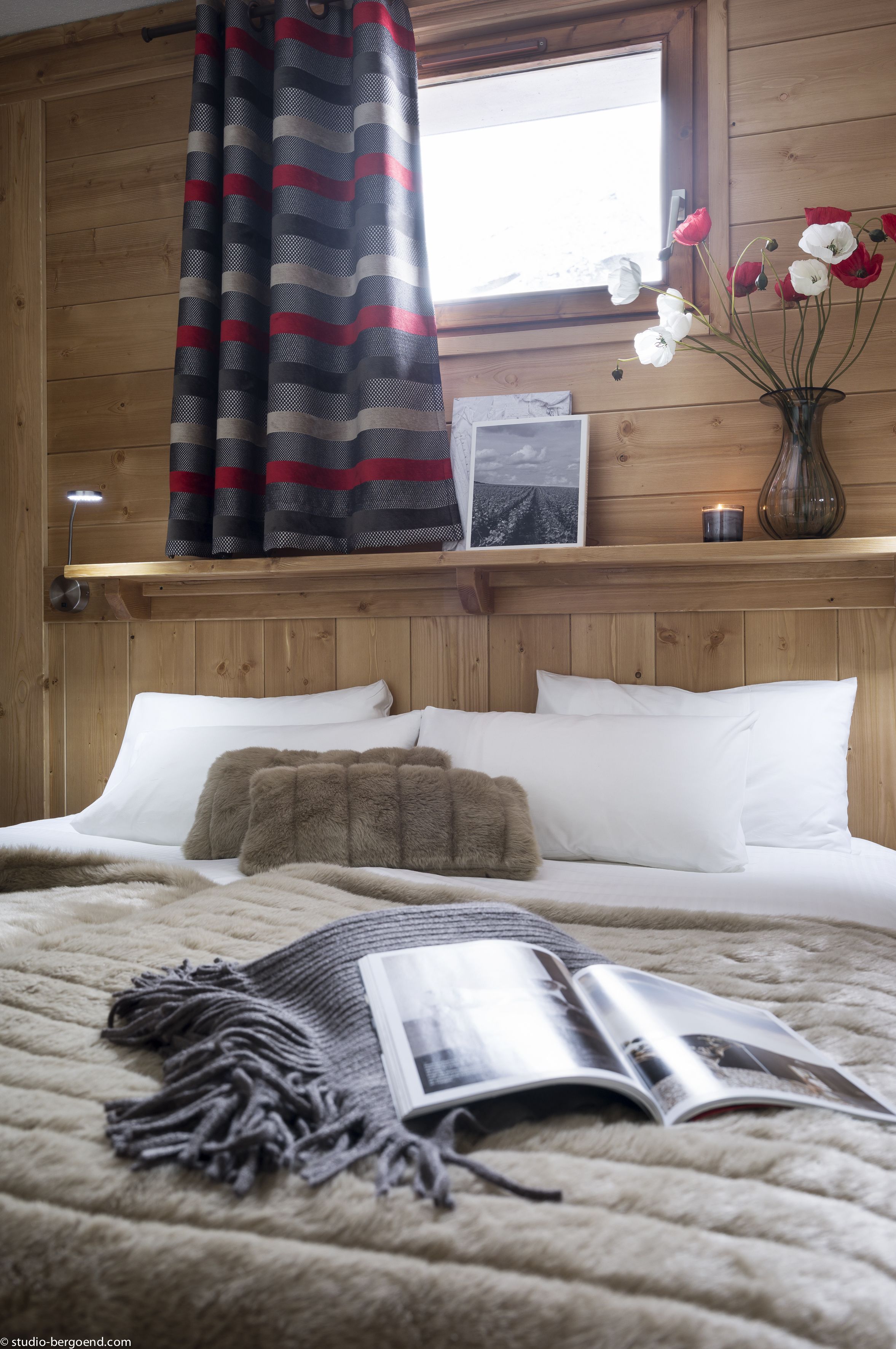 Un dormitorio con cabecera de madera, una ventana con cortinas a rayas, una mesita de noche con una vela y un jarrón con flores.
