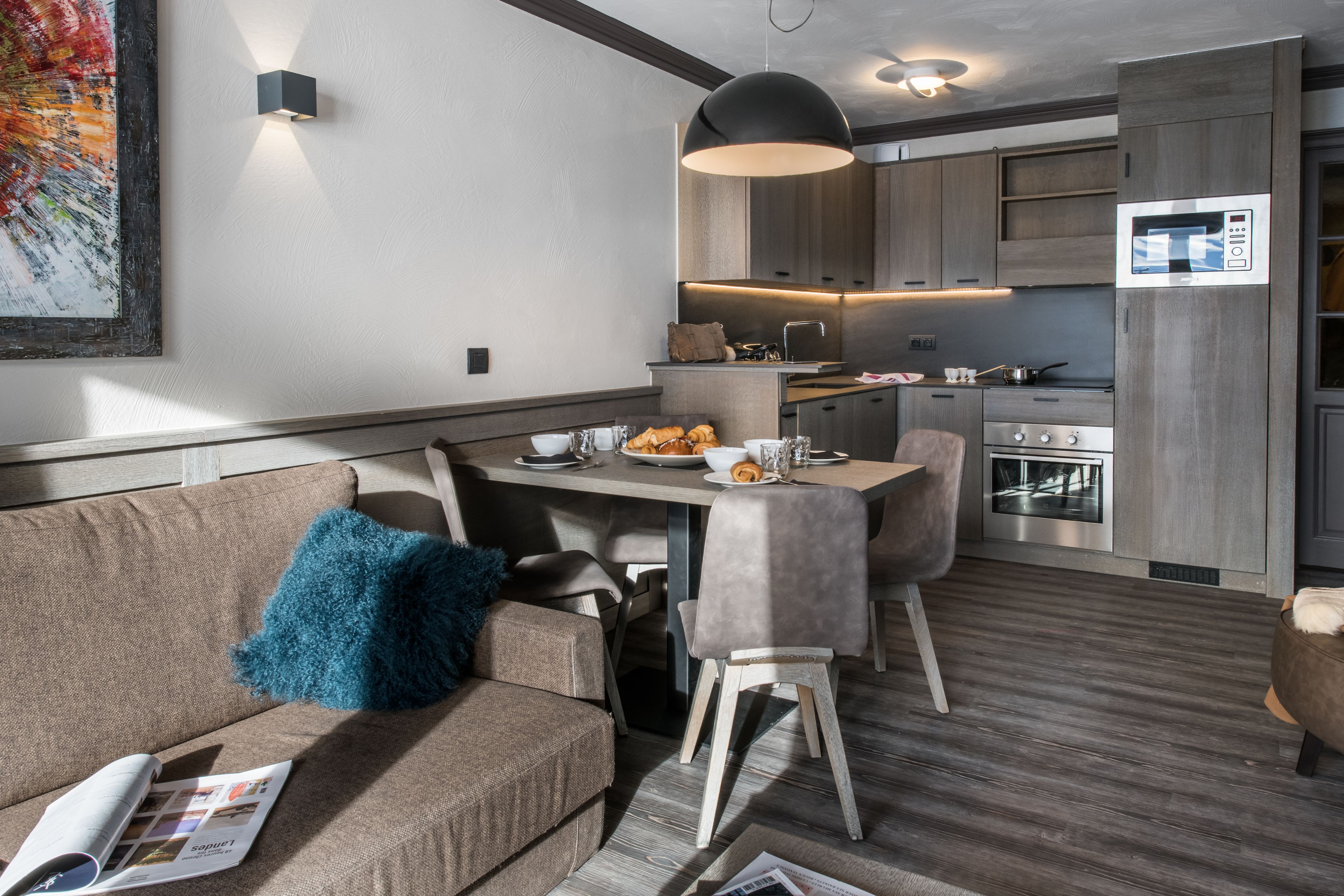 A modern kitchen with a dining area, featuring a wooden floor, a sofa with a blue pillow, and a table set for two.
