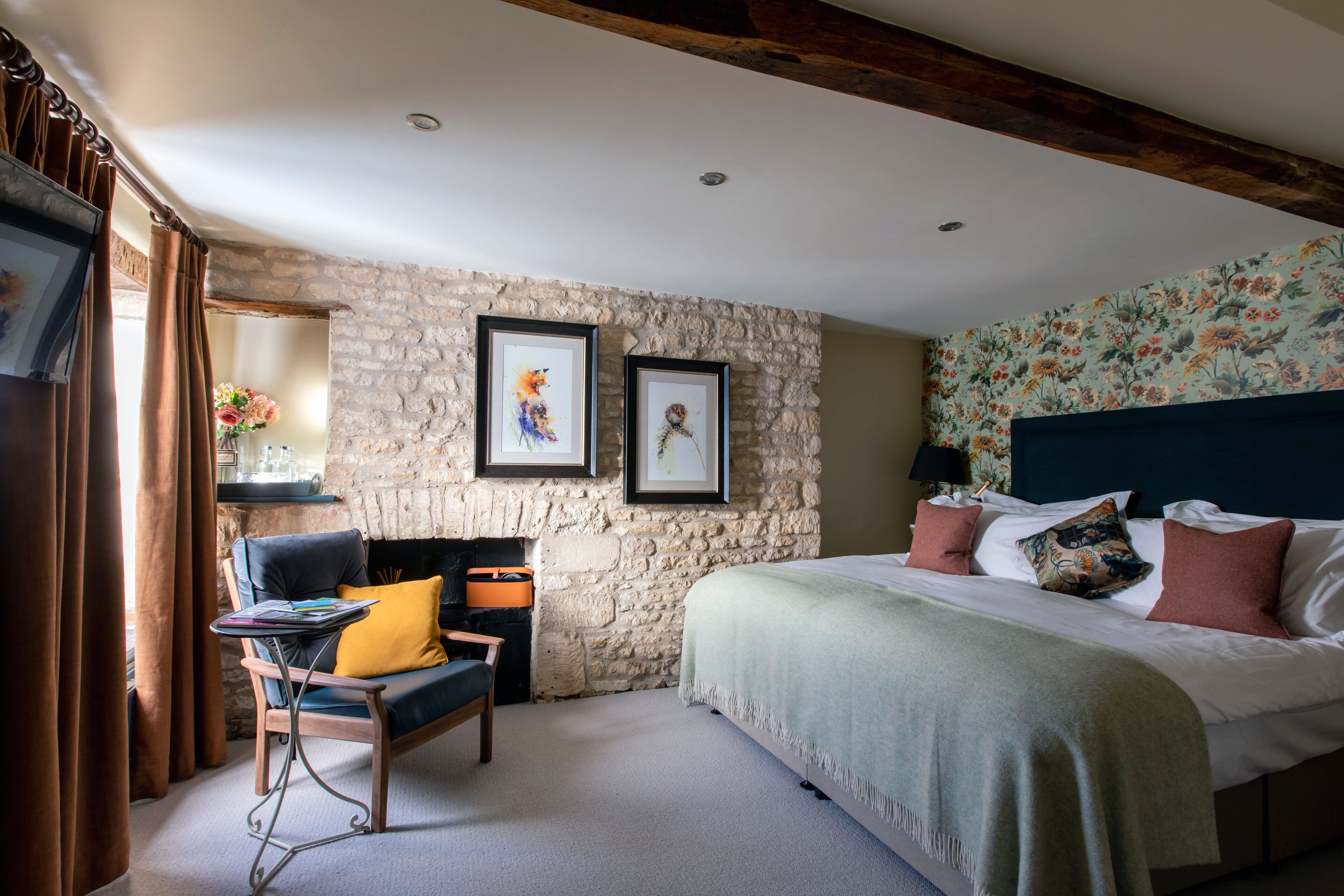 A bedroom with a stone wall, a bed with multiple pillows, a chair with a yellow cushion, and a small table with books.