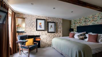 A bedroom with a stone wall, a bed with multiple pillows, a chair with a yellow cushion, and a small table with books.