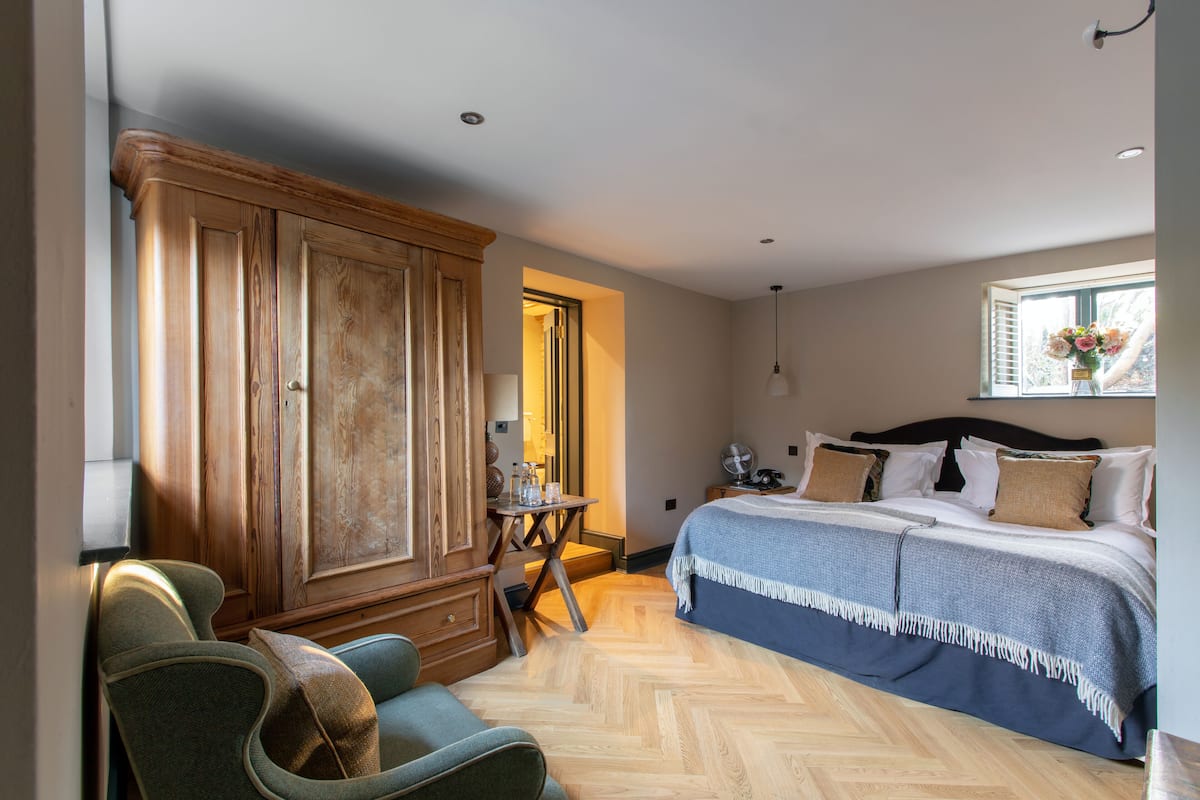 A bedroom with a large wooden wardrobe, a bed with a blue blanket, a small table with a lamp, and a window with a view of trees.