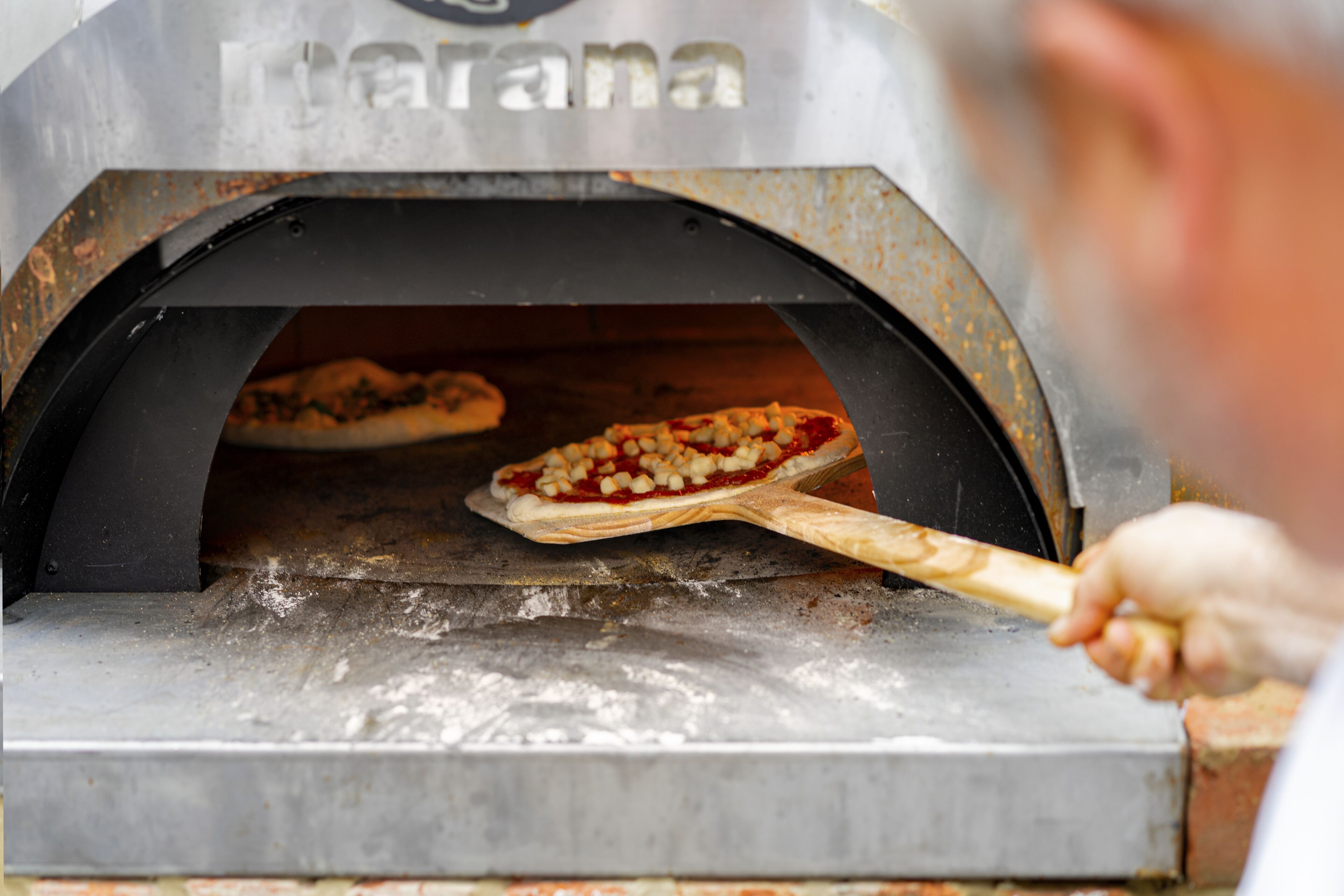 A pizza oven with two pizzas inside, one being removed with a wooden pizza peel.