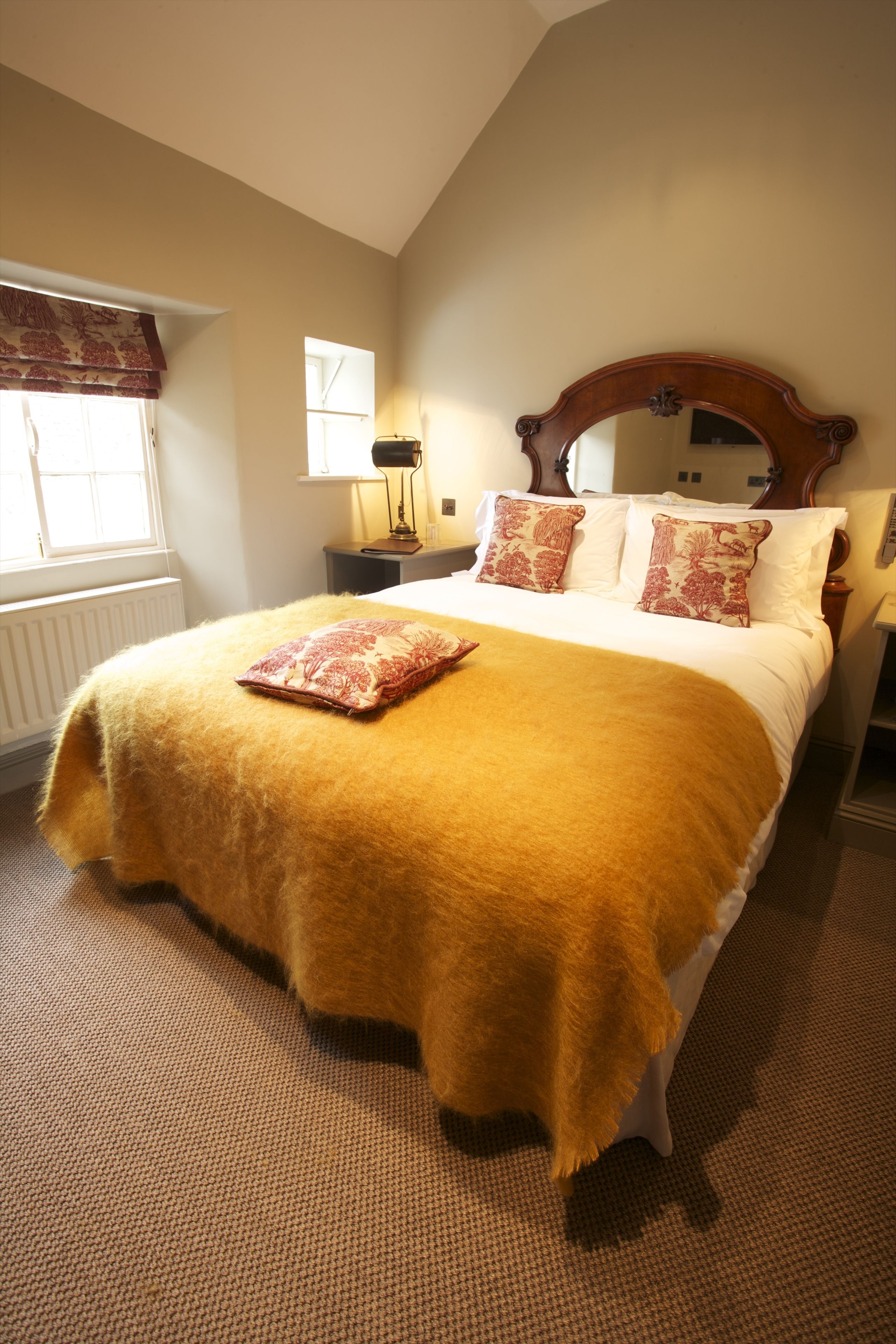 A bedroom with a large bed, a wooden headboard, a window with curtains, a bedside table with a lamp, and a mirror on the wall.