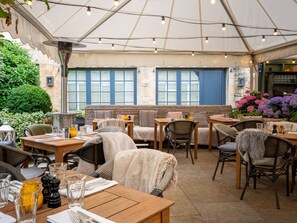 An outdoor dining area with tables and chairs, a covered section with string lights, and a view of the street with buildings and a blue door. - The Wheatsheaf Inn (Cheltenham)