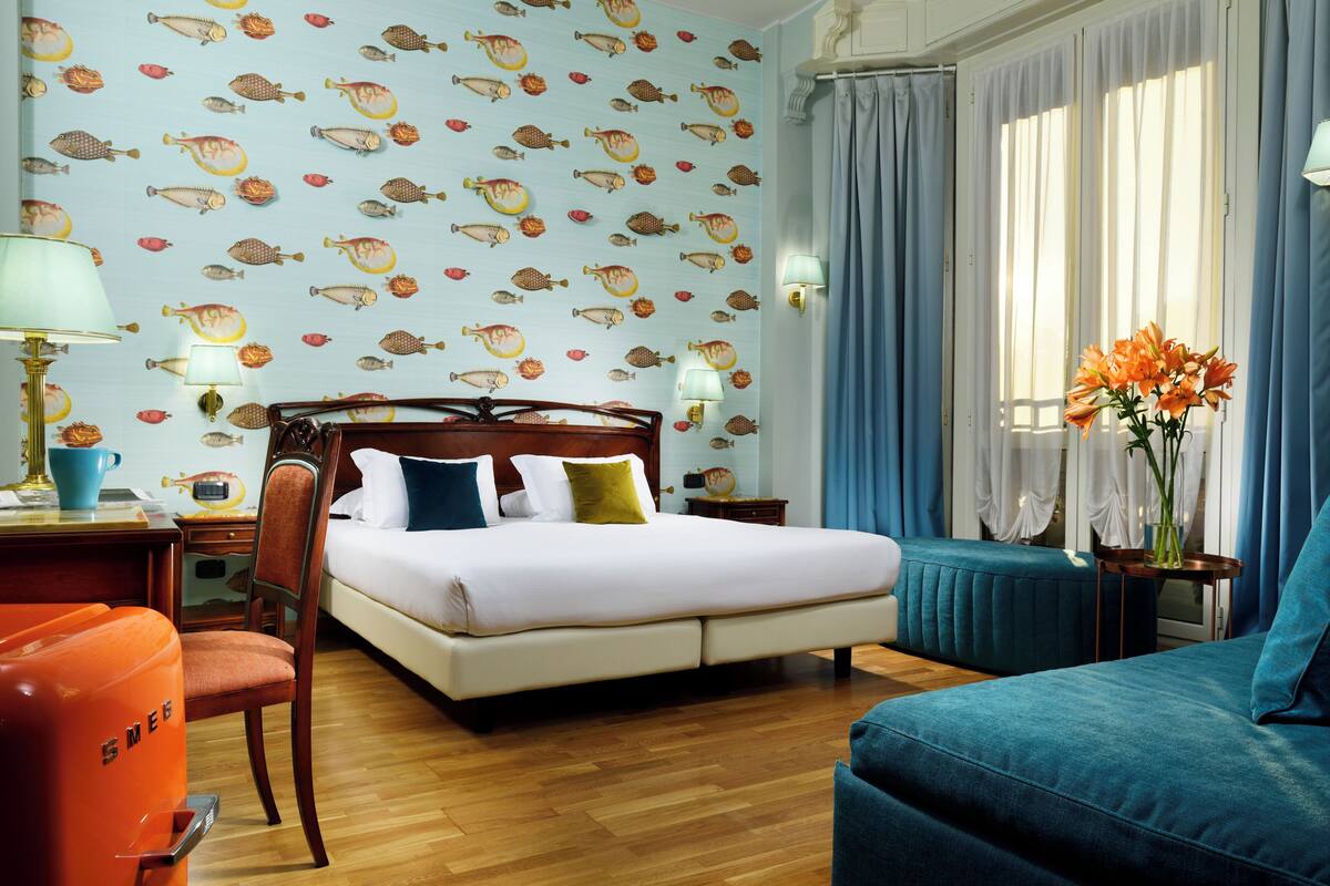 Una habitación de hotel con cama, una silla, un escritorio con lámpara, una mesita de noche y una ventana con cortinas.
