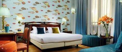 A hotel room with a bed, a chair, a desk with a lamp, a nightstand, and a window with curtains.