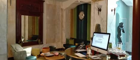 A modern living room with a round desk, a computer, and a statue. There are two chairs, a sofa, and a coffee table. The room has a marble floor and a large mirror.