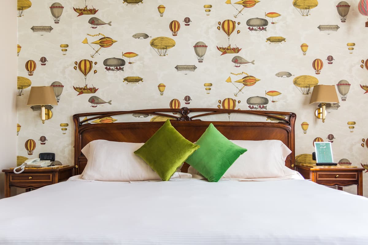 Habitación de hotel con cama, dos almohadas verdes, cabecera de madera, mesita de noche con lámpara y pared con papel tapiz de globo aerostático.