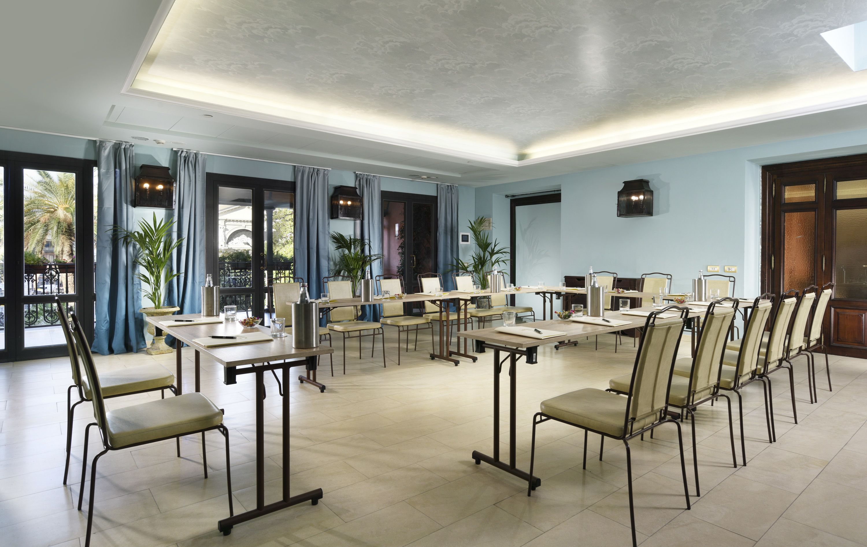 Une salle de conférence dotée de plusieurs tables et chaises, de grandes fenêtres avec des rideaux et d’un plafond équipé d’un éclairage encastré.