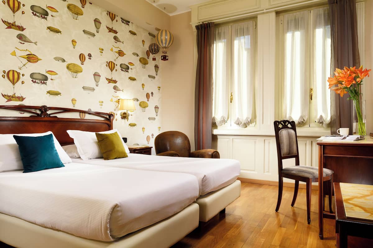 Habitación de hotel con dos camas, cabecero de madera, escritorio con silla, ventana con cortinas y pared con papel tapiz de motivos de globos.
