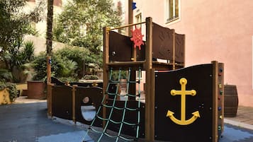 A children's play structure with a rope net and anchor design, set against a building with a pink facade.