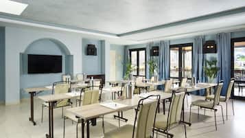 Uma sala de conferências com várias mesas e cadeiras, uma TV de tela plana na parede e grandes janelas com cortinas.