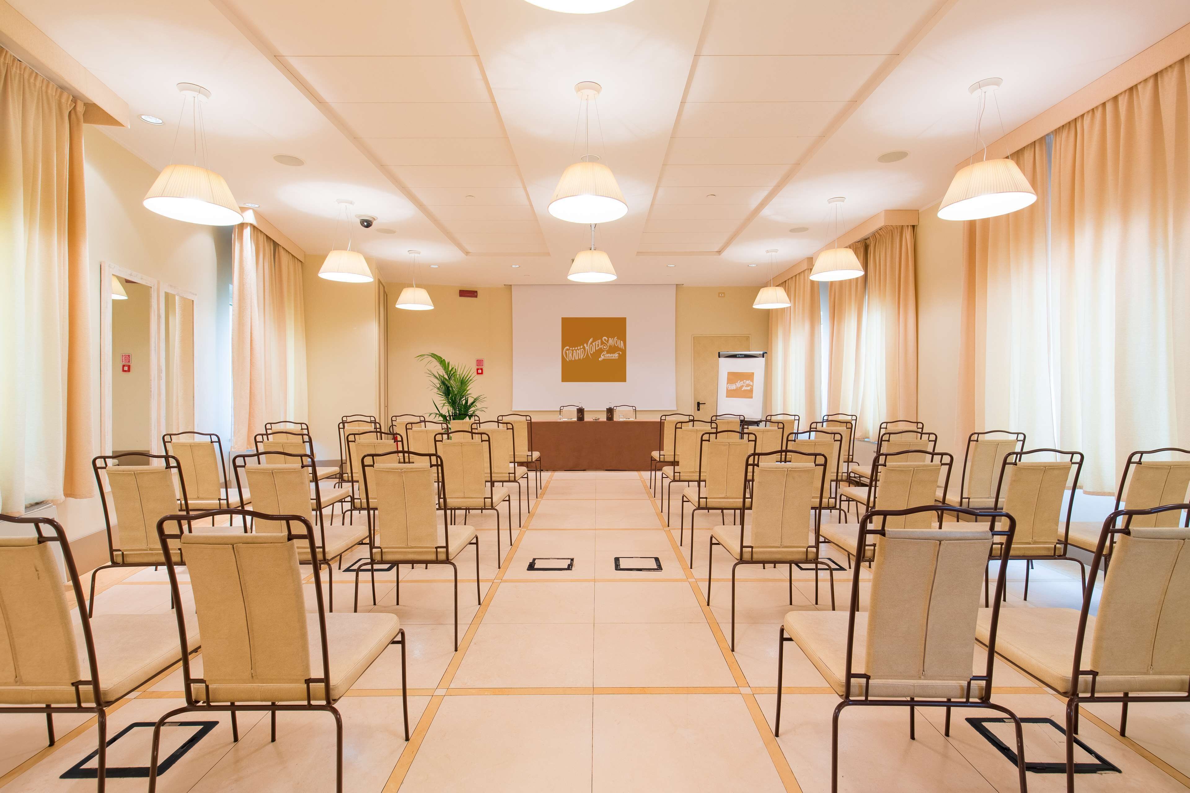 Ein Konferenzraum mit Reihen von Stühlen, die zu einem Rednerpult blicken, sowie beigefarbenen Vorhängen und Deckenleuchten.