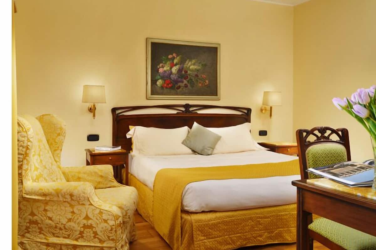 Una habitación de hotel con cama, una silla, un escritorio, una lámpara y un cuadro en la pared.