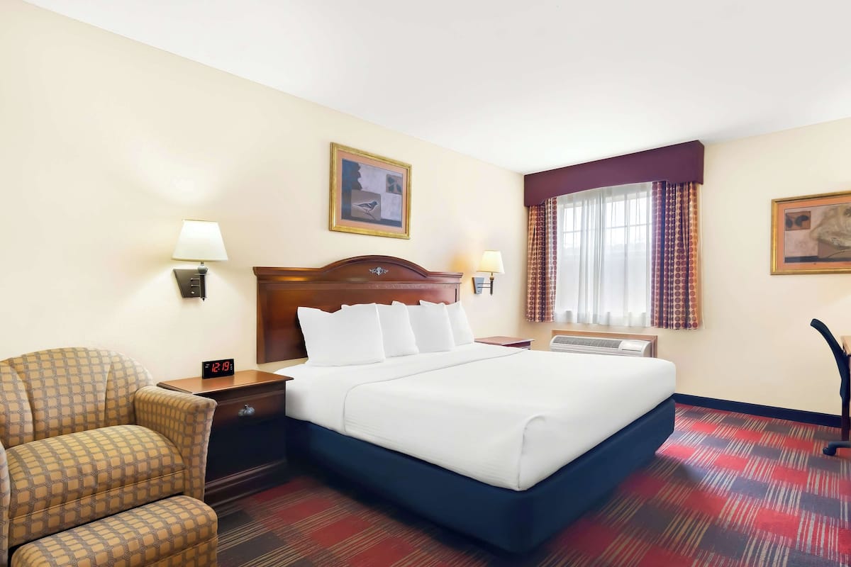 Een hotelkamer met een bed, een stoel, een nachtkastje, een lamp, een klok en een raam met gordijnen.