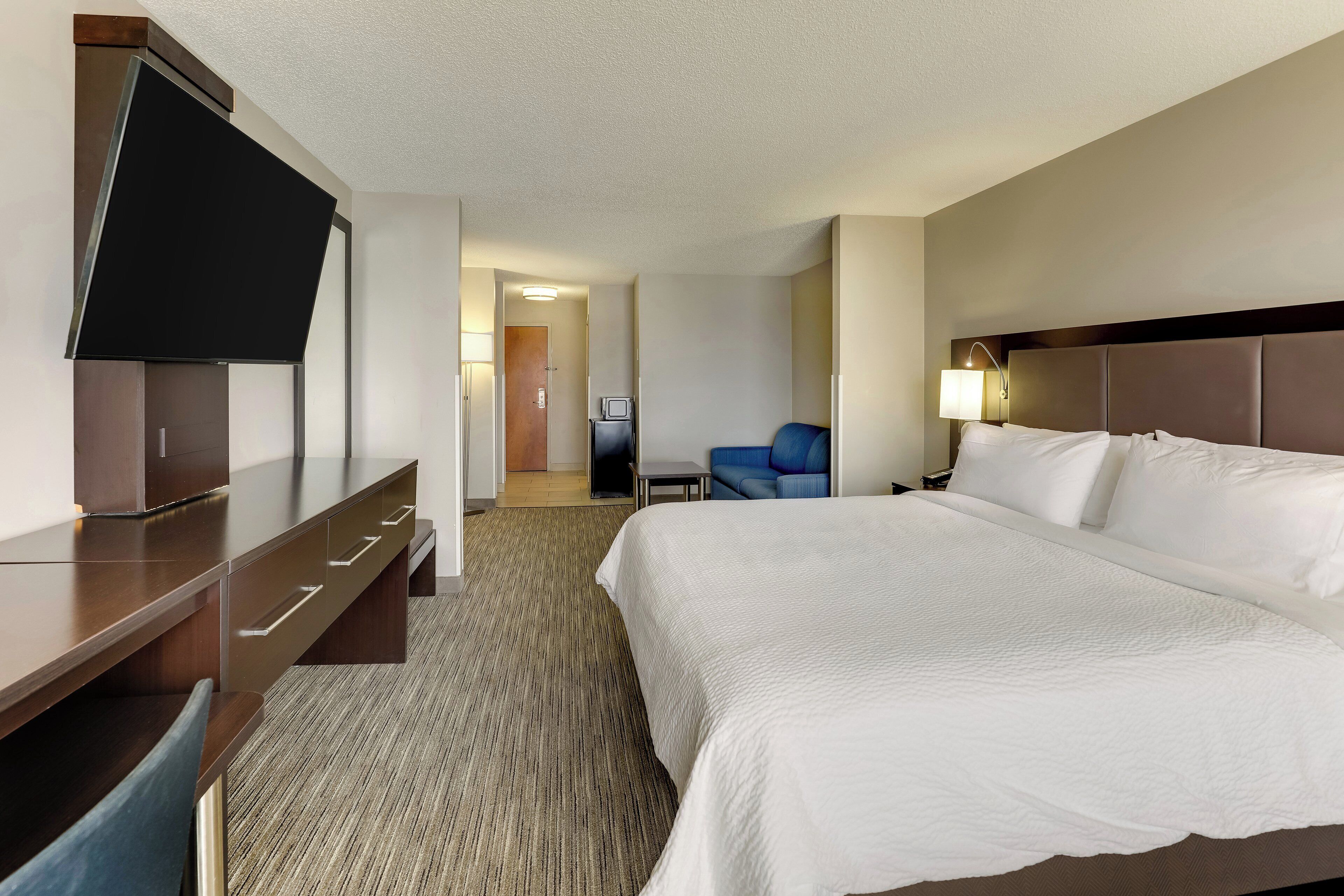A hotel room with a large bed, a flat-screen TV, a desk with drawers, a chair, and a small table.
