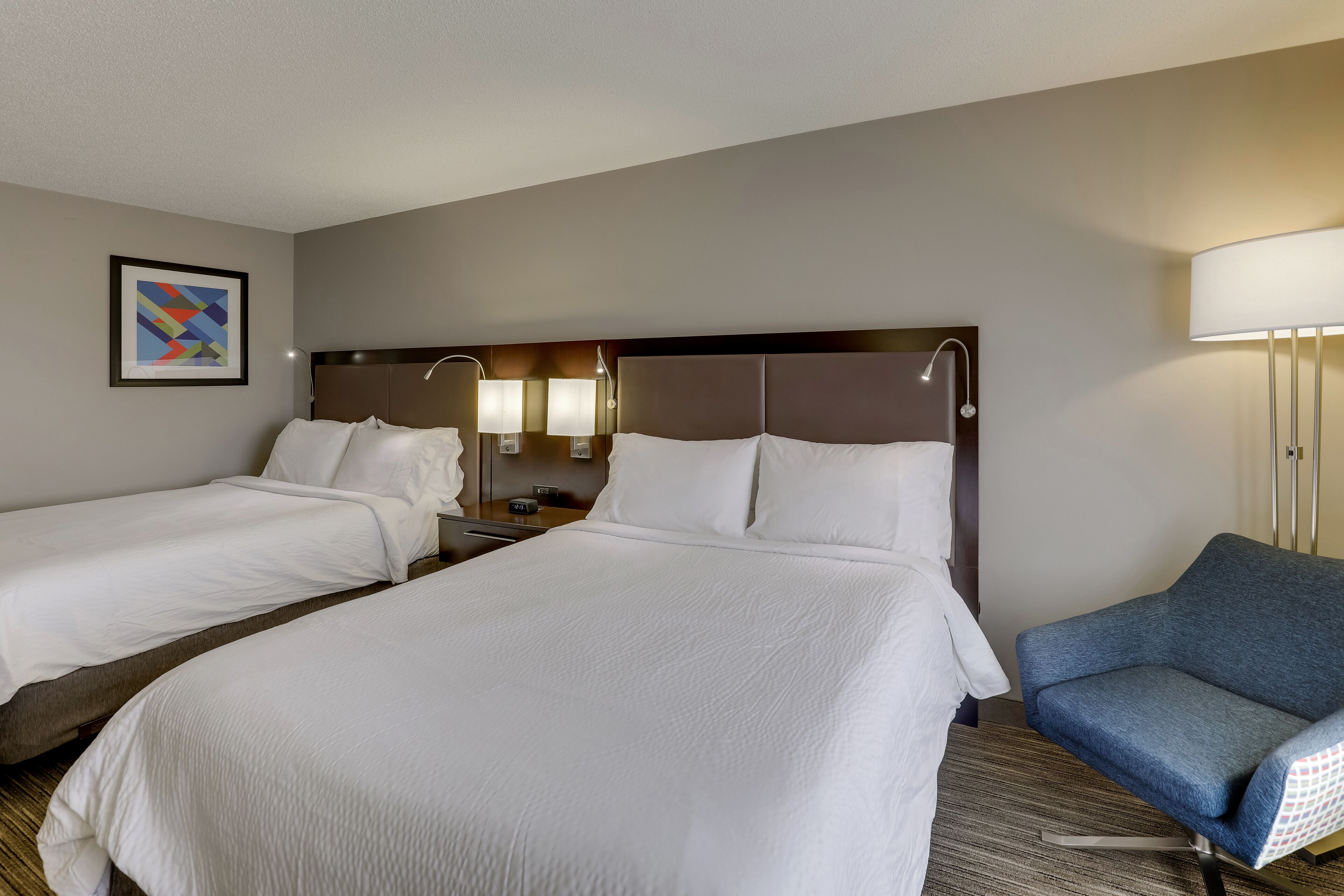 A hotel room with two beds, a nightstand, a lamp, and a blue armchair.
