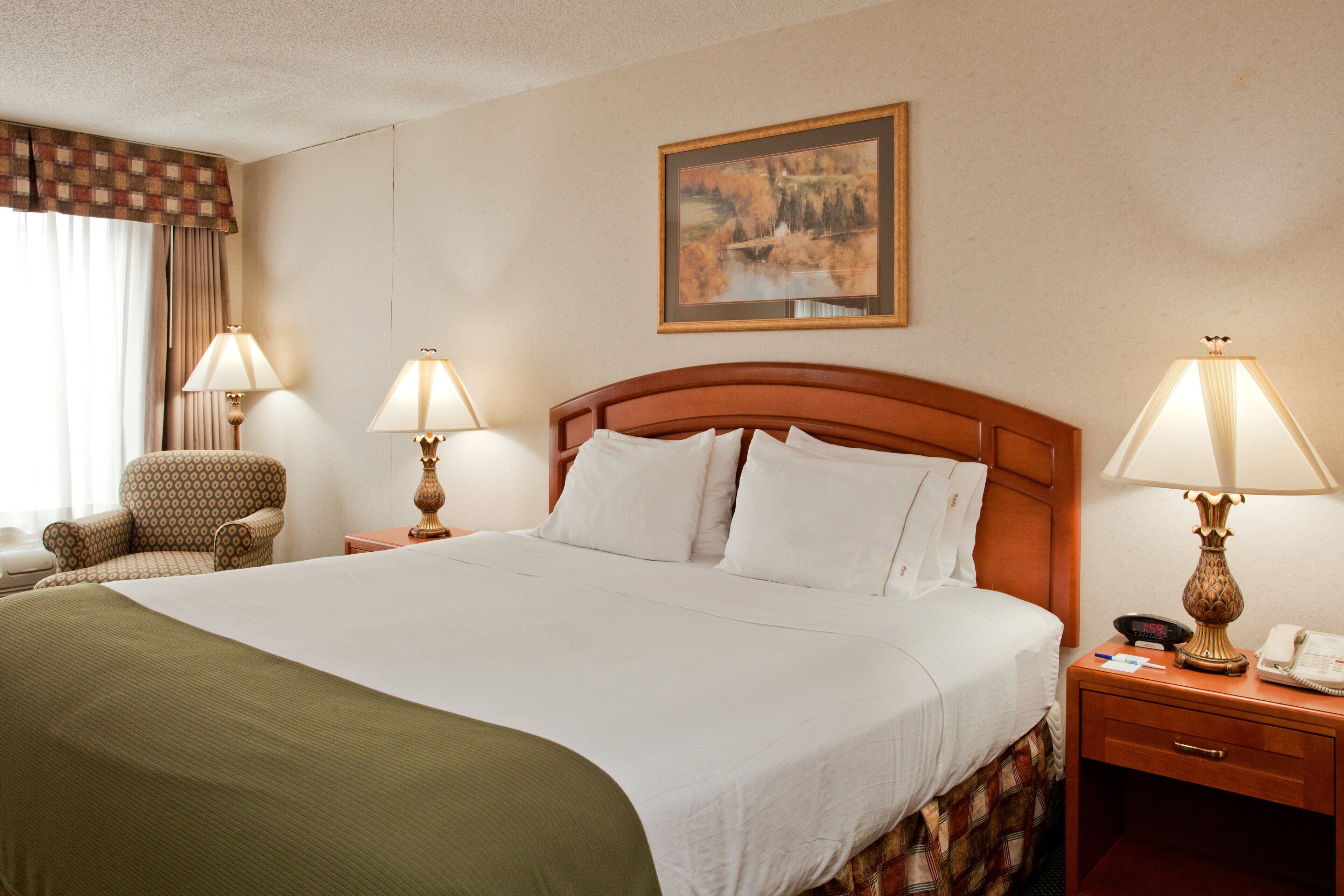 A hotel room with a large bed, two bedside lamps, a chair, a desk with a telephone, and a painting on the wall.