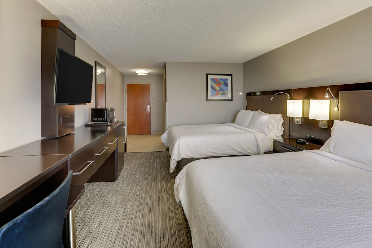 Quarto de hotel com duas camas, uma escrivaninha com televisão e porta de banheiro.