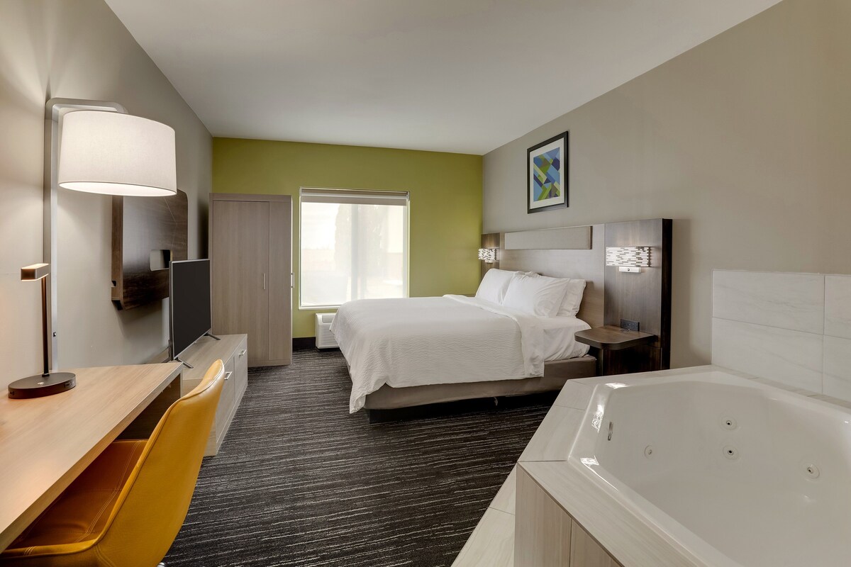 Quarto de hotel com cama, escrivaninha, cadeira, luminária de mesa, mesa de cabeceira, televisão, janela e banheira.