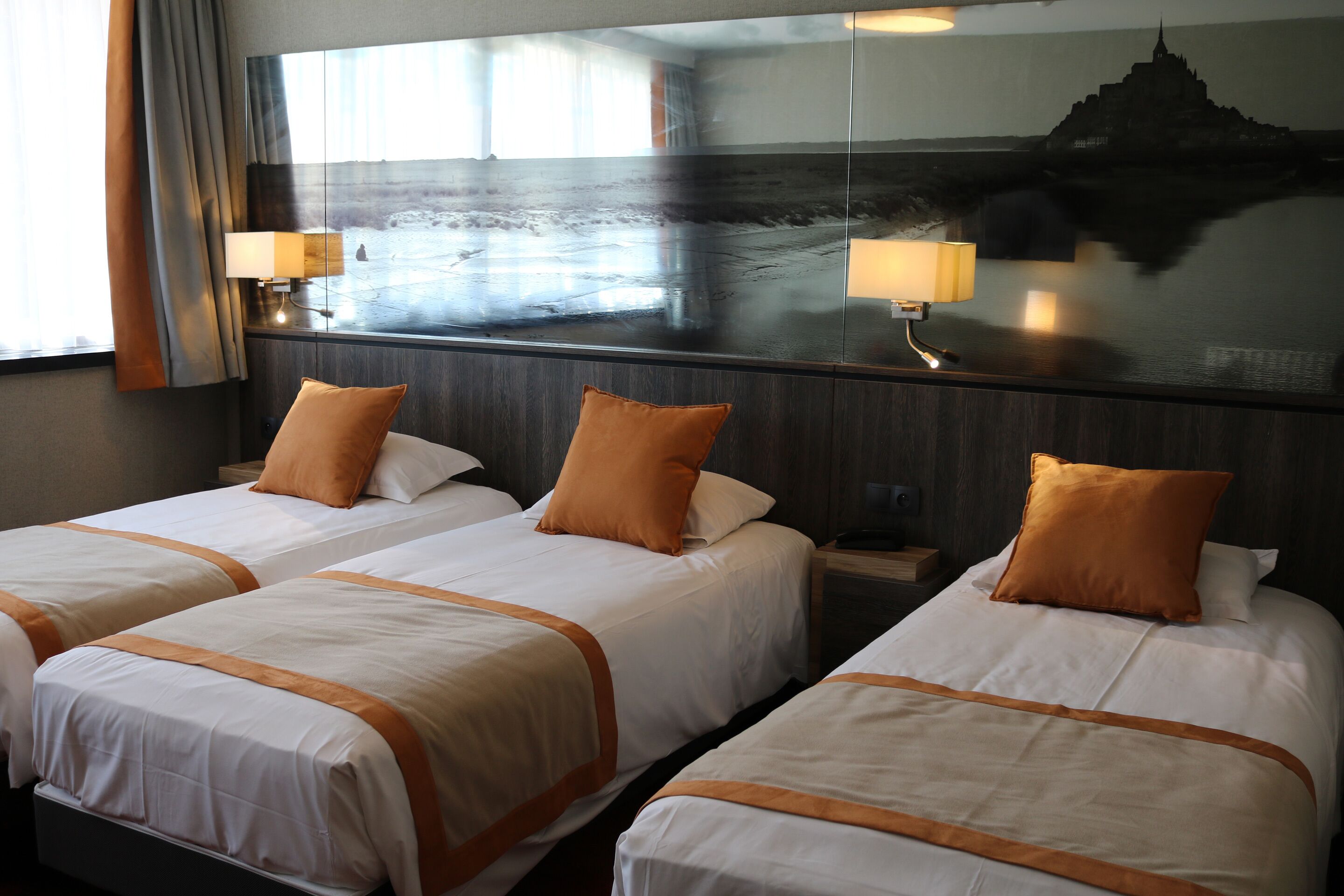 A hotel room with two beds, each with a white bedspread and orange accents, a large mirror reflecting a view of a temple, and a nightstand with a lamp.