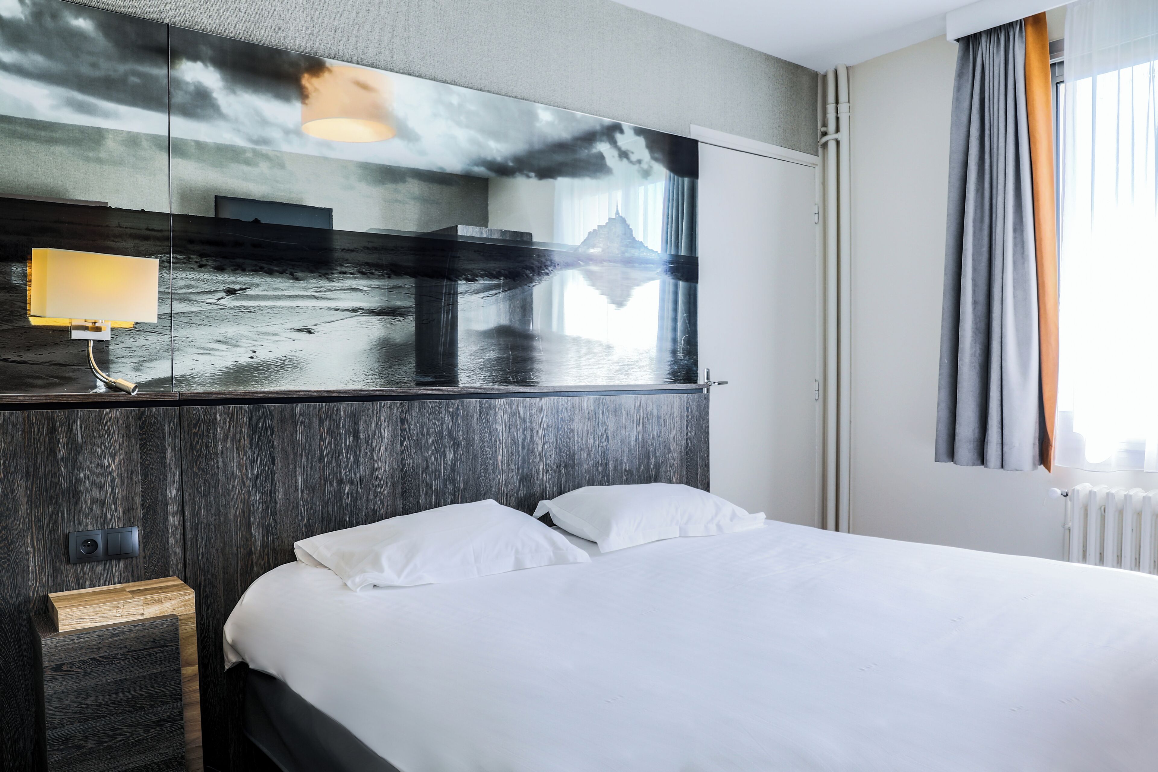 Une chambre d’hôtel avec un grand lit, une table de chevet, une lampe et un miroir mural qui reflète un paysage magnifique.