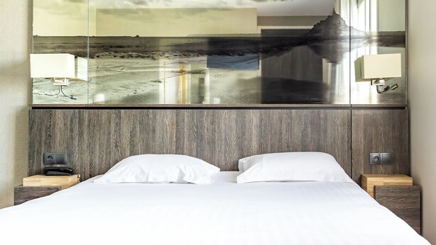 A hotel room with a large mirror reflecting a beach scene, a wooden headboard, two bedside tables with lamps, and a neatly made bed. - Hôtel de la Digue (Le Mont-Saint-Michel)