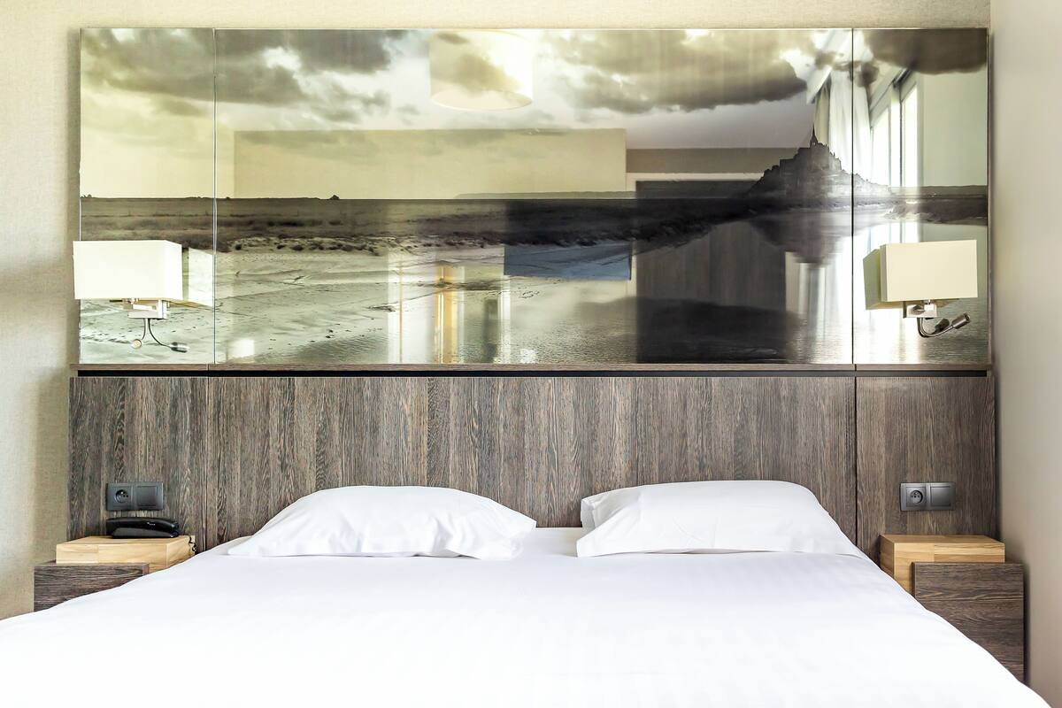 A hotel room with a large mirror reflecting a beach scene, a wooden headboard, two bedside tables with lamps, and a neatly made bed.