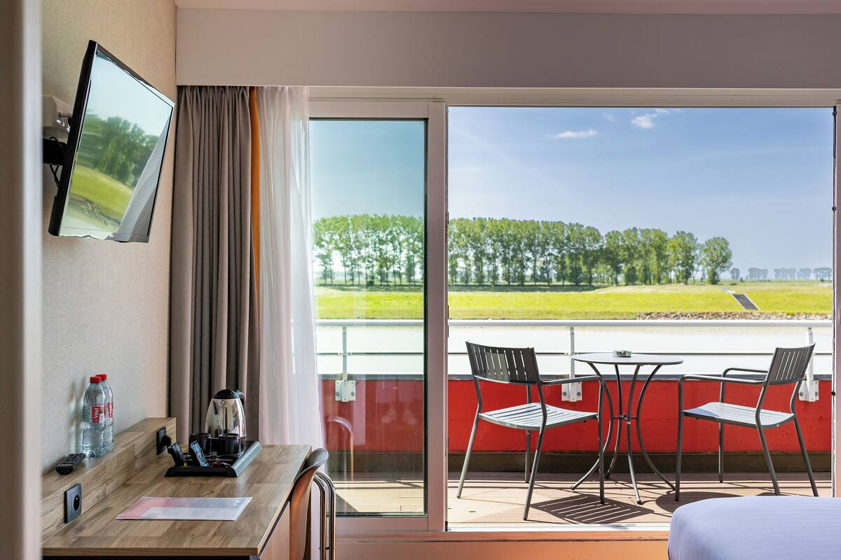 A hotel room with a balcony, a desk with a coffee maker, a TV, and a view of a green landscape.
