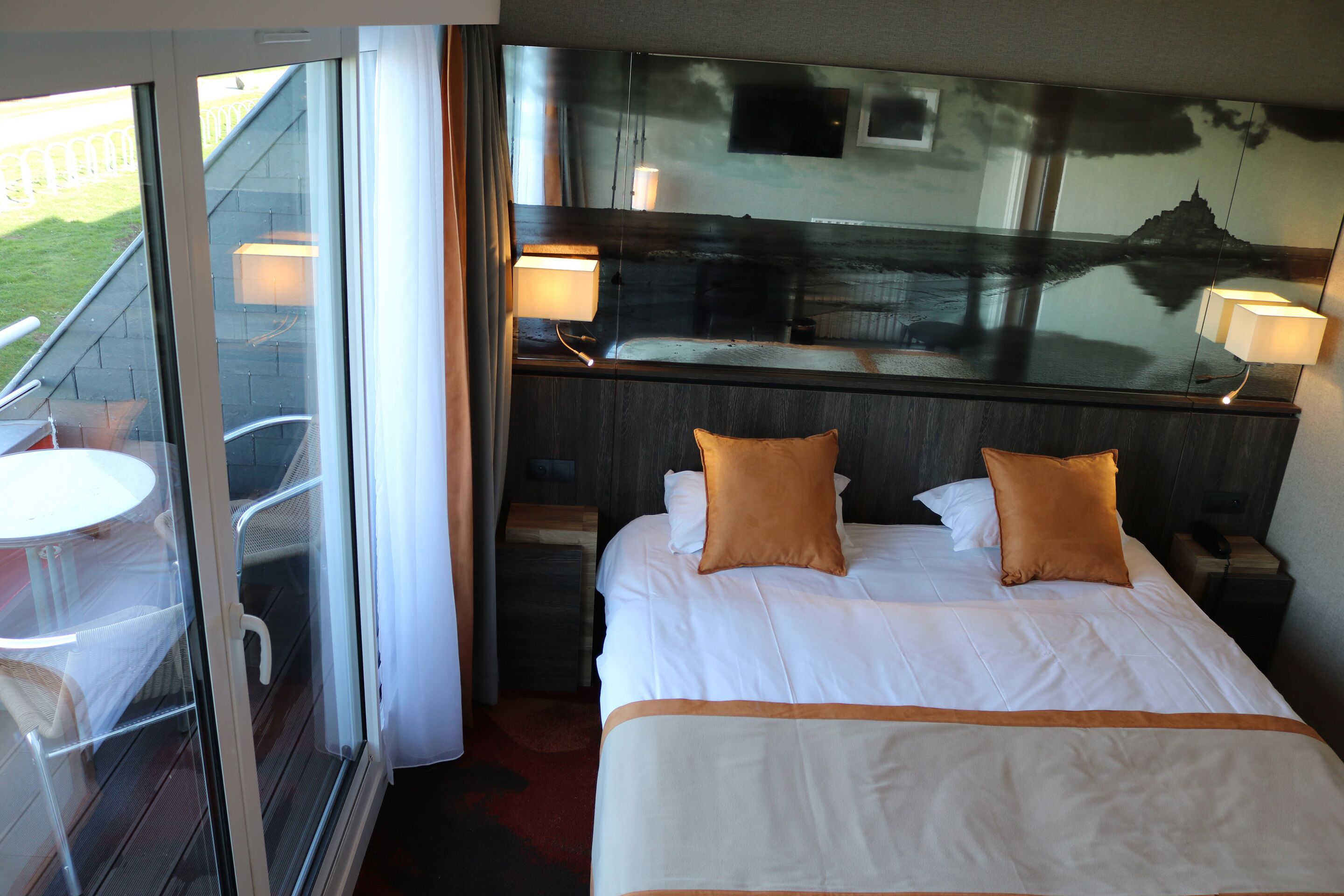 Habitación de hotel con una cama, dos almohadas, una mesita de noche, una lámpara y vistas al exterior a través de un ventanal.