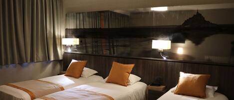 A hotel room with three beds, each with white bedding and orange accents. There is a large mirror on the wall, and a nightstand with a lamp.