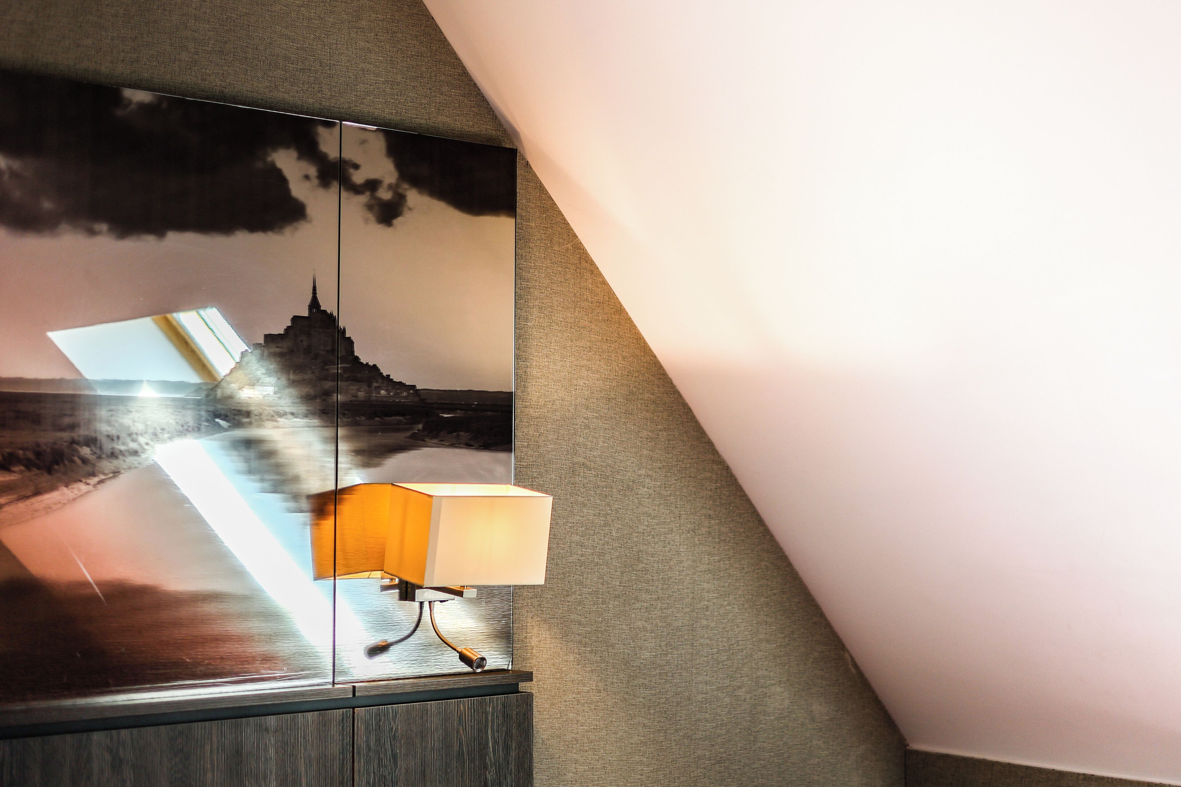 A room with a large mirror reflecting a cityscape, a lamp on a wooden cabinet, and a textured wall.