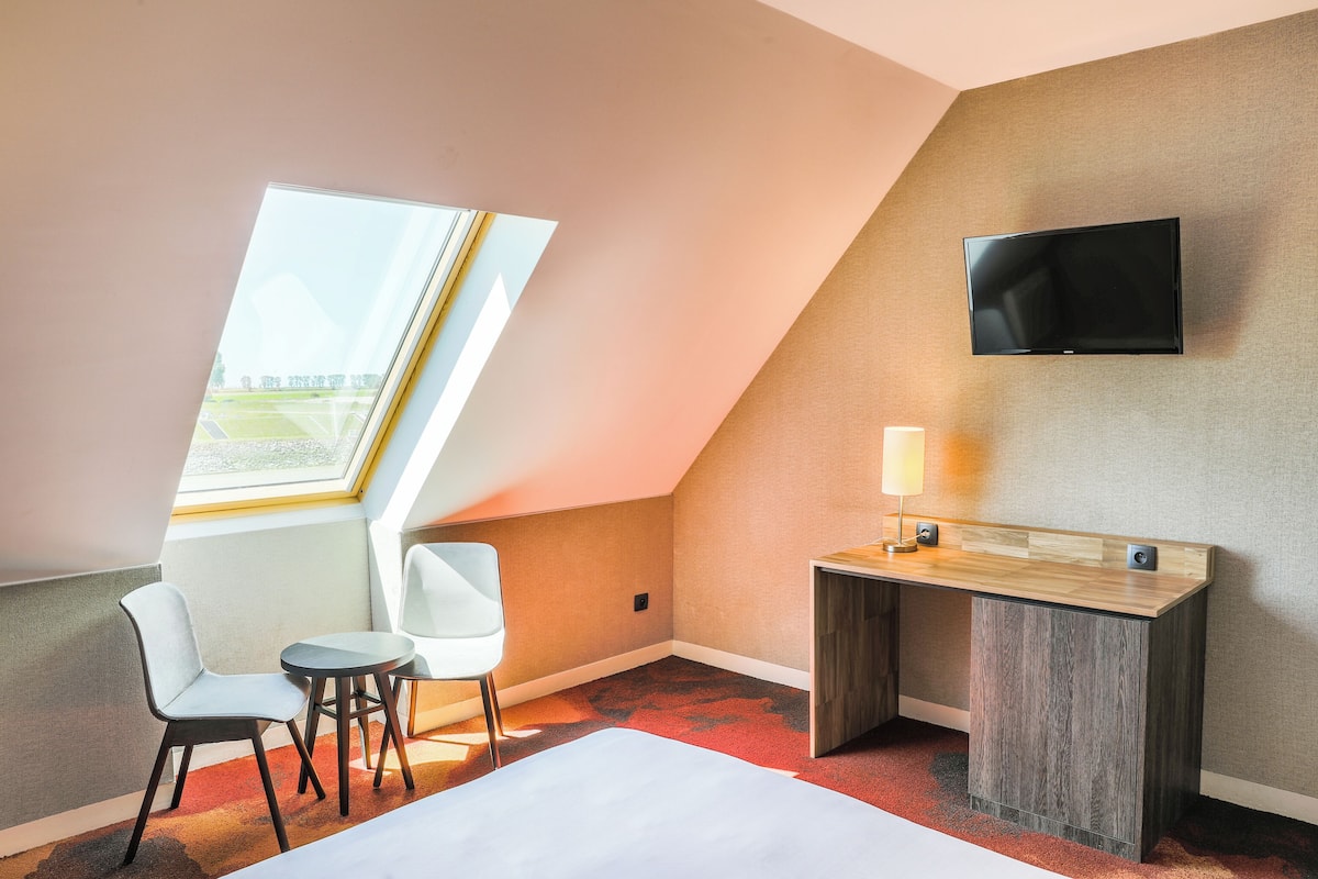 A hotel room with a sloped ceiling, a flat-screen TV mounted on the wall, a desk with a lamp, a bed, and two chairs.