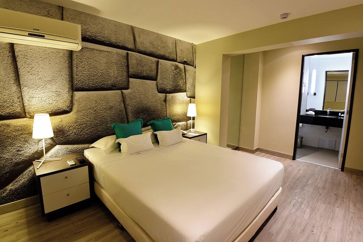 A hotel room with a large bed, two bedside tables with lamps, a stone-textured headboard, and a view of the bathroom through an open door.