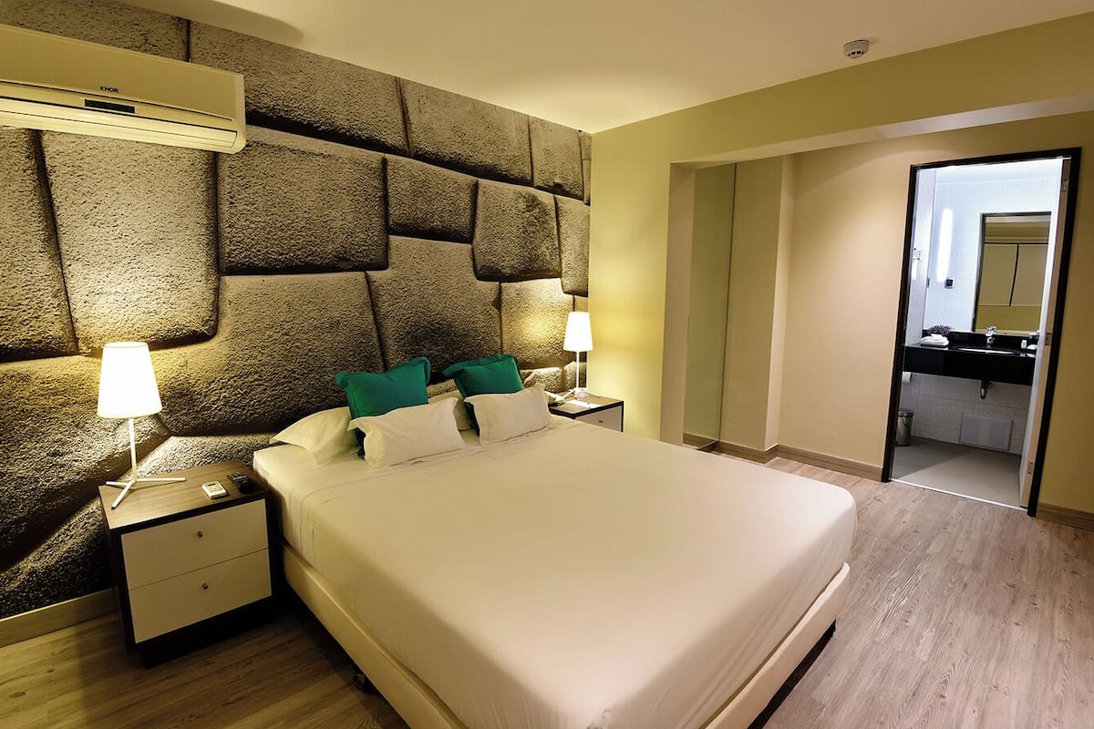 Un cuarto de hotel con una cama grande, dos mesitas de noche con lámparas, un cabecero de textura pétrea y vista al baño a través de una puerta abierta.