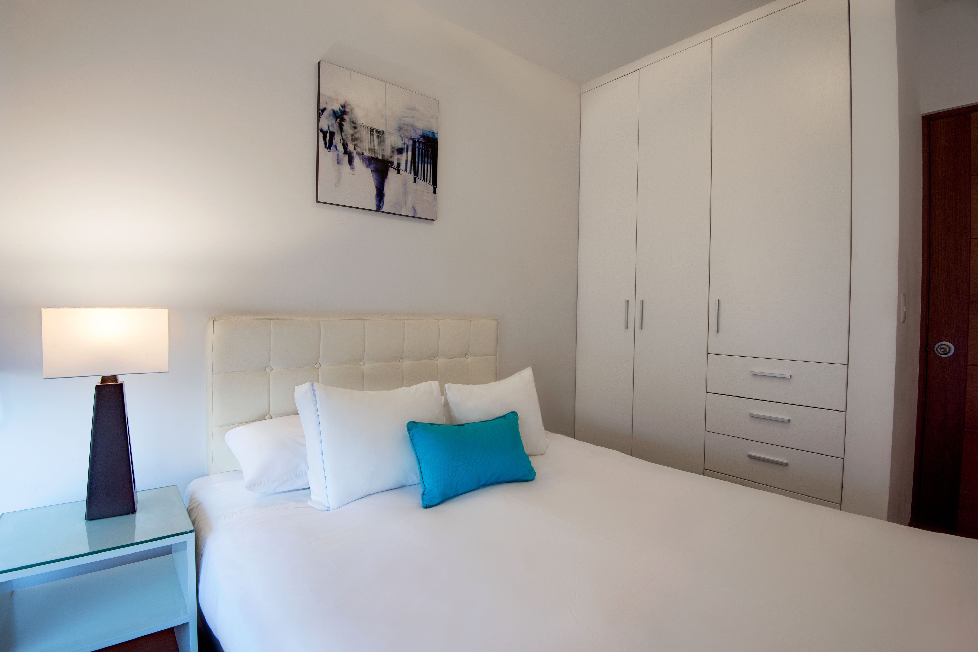 Un dormitorio con cama, mesita de noche, lámpara, cabecera blanca, almohada azul, pared blanca, un cuadro y un armario.