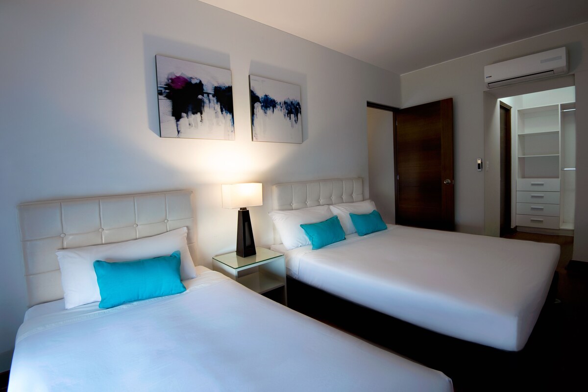 A hotel room with two beds, a nightstand with a lamp, and artwork on the wall.