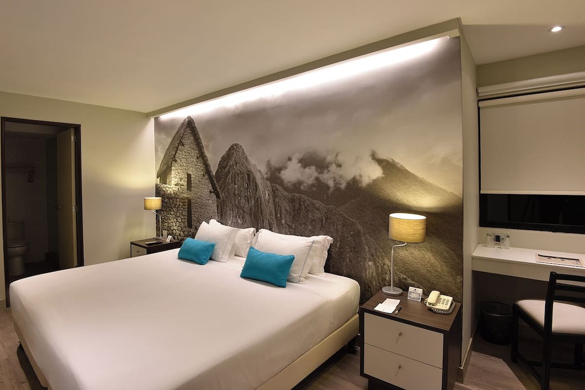 A hotel room with a large bed, a desk with a chair, a telephone, and a wall-mounted lamp.