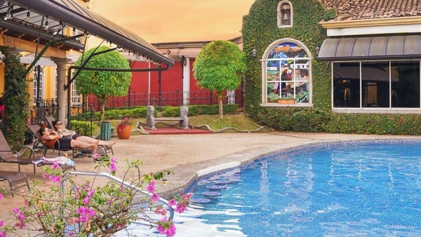 Ein Poolbereich mit Liegestühlen, einer überdachten Sitzgelegenheit und einem Gebäude mit einer grünen, efeuüberrankten Wand und einem Fenster mit Blick auf den Pool.
