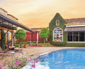 A swimming pool area with lounge chairs, a covered seating area, and a building with a green ivy-covered wall and a window with a view of the pool. - Casa Conde Apartamentos (San José)