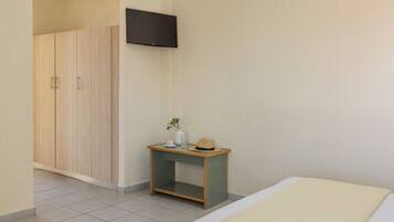 Un dormitorio con una cama, un televisor en la pared, un armario de madera y una mesita con un sombrero y un florero.