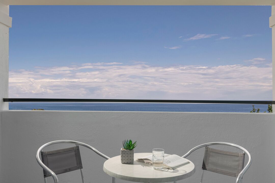 En balkong med ett runt bord, två stolar, en liten krukväxt och ett glas vatten.