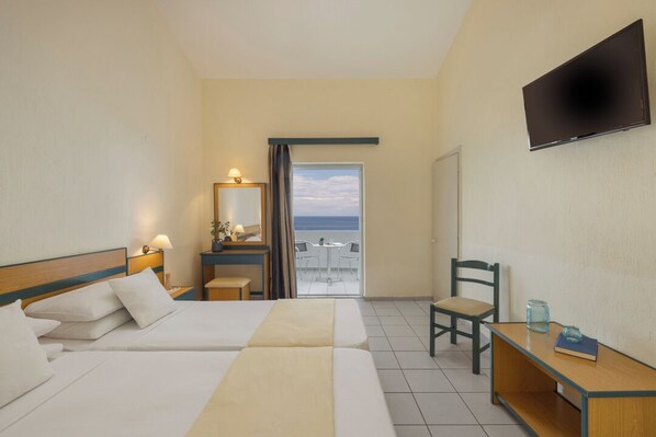 A hotel room with two beds, a desk, a chair, a TV, and a balcony with a view. - Porto Skala Hotel Village (Kefalonia)
